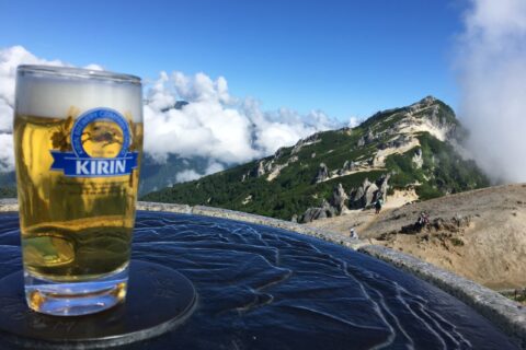 雲海の向こうに連なる山の稜線を眺めながら、山小屋のテラスでビールを楽しむ登山後の風景